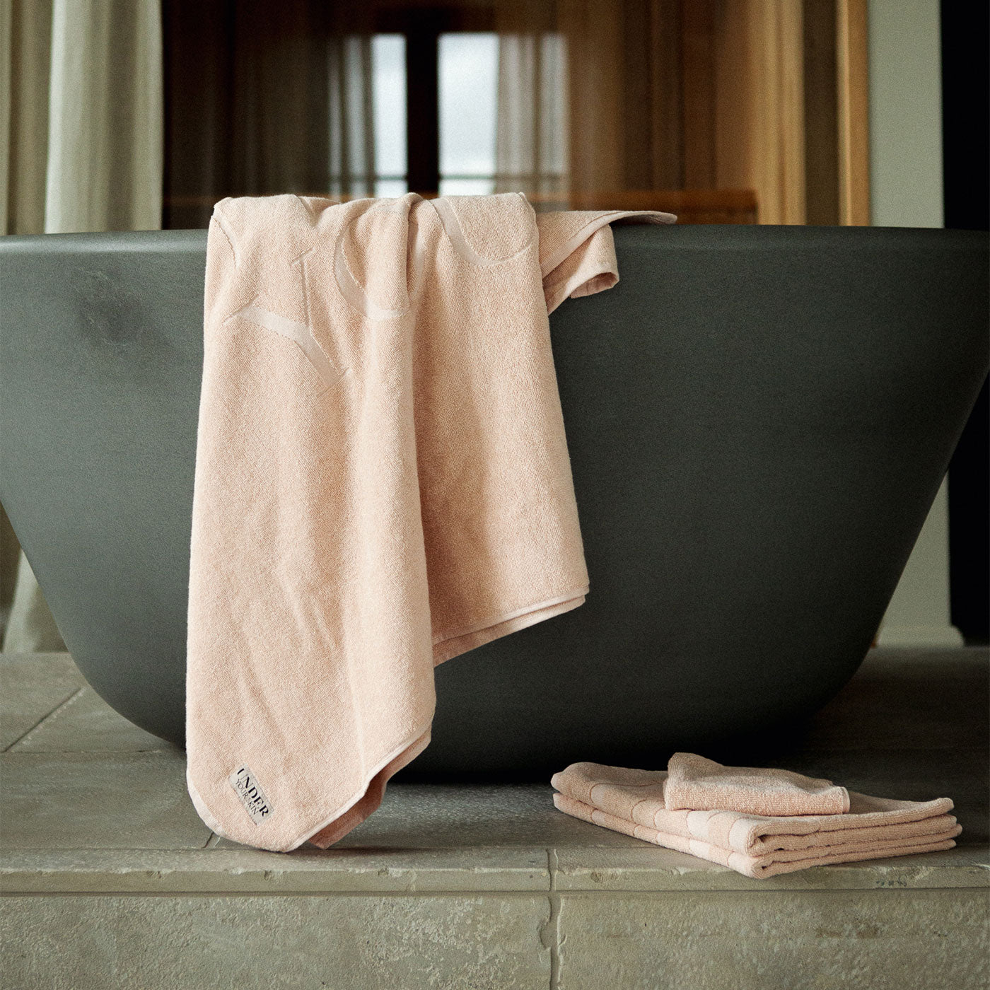 Rosa badhandduk över badkarskanten i badrumsmiljö, med två handdukar framför badkaret.