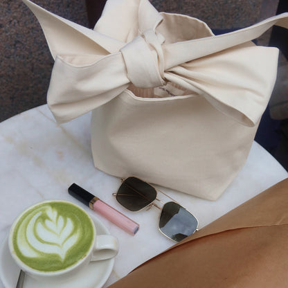 Bow bag på ett bord i cafémiljö tillsammans med en matcha latte, ett läppstift och solglasögon.