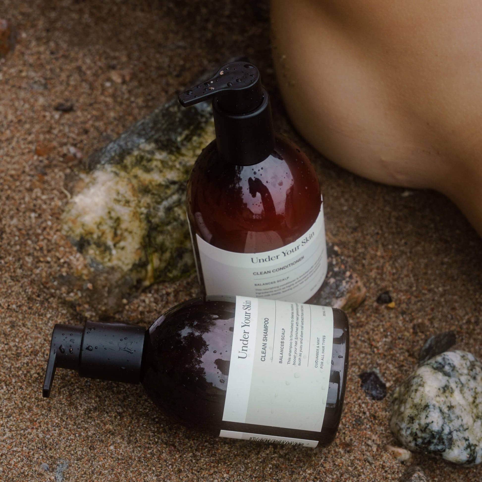 Hårvårdsprodukter placerade på sandstrand med stenar intill och en fot bredvid.