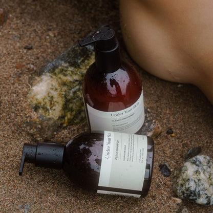 Hårvårdsprodukter placerade på sandstrand med stenar intill och en fot bredvid.