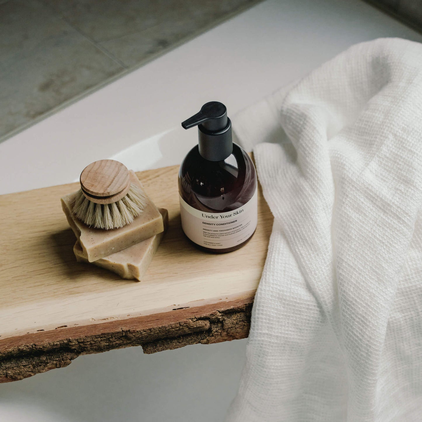 Flaska Under Your Skin Density Conditioner på en träplanka över ett badkar, tillsammans med en träborste och vikt handduk.