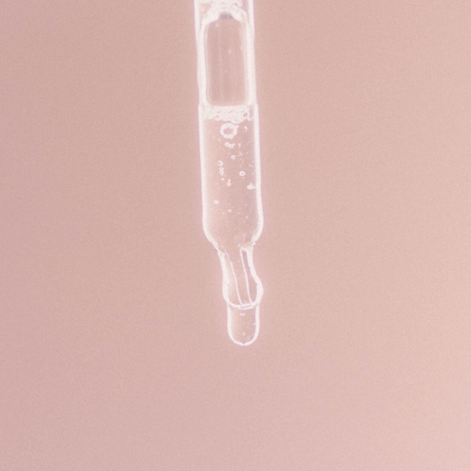 Närbild på en glas­pipett med klar vätska mot en mjuk rosa bakgrund