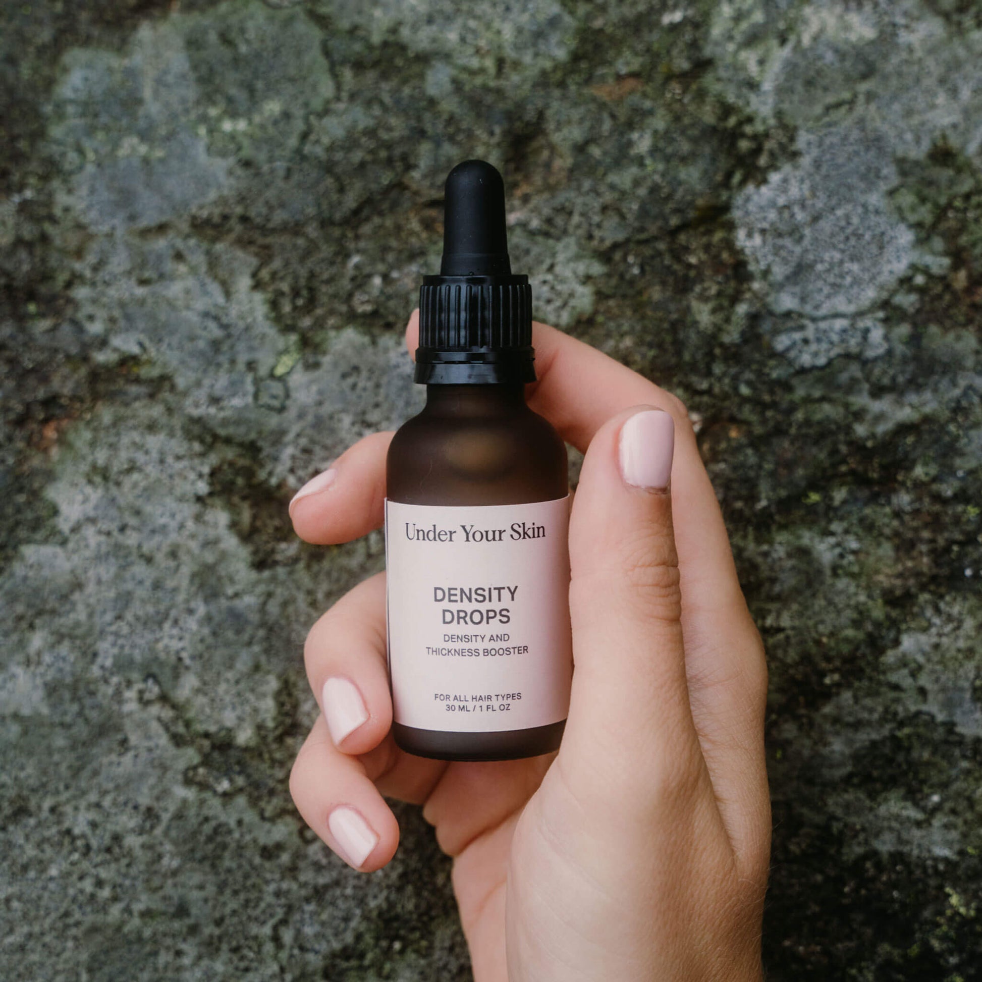 Hand som håller en flaska Under Your Skin Density Drops framför en texturerad stenyta.