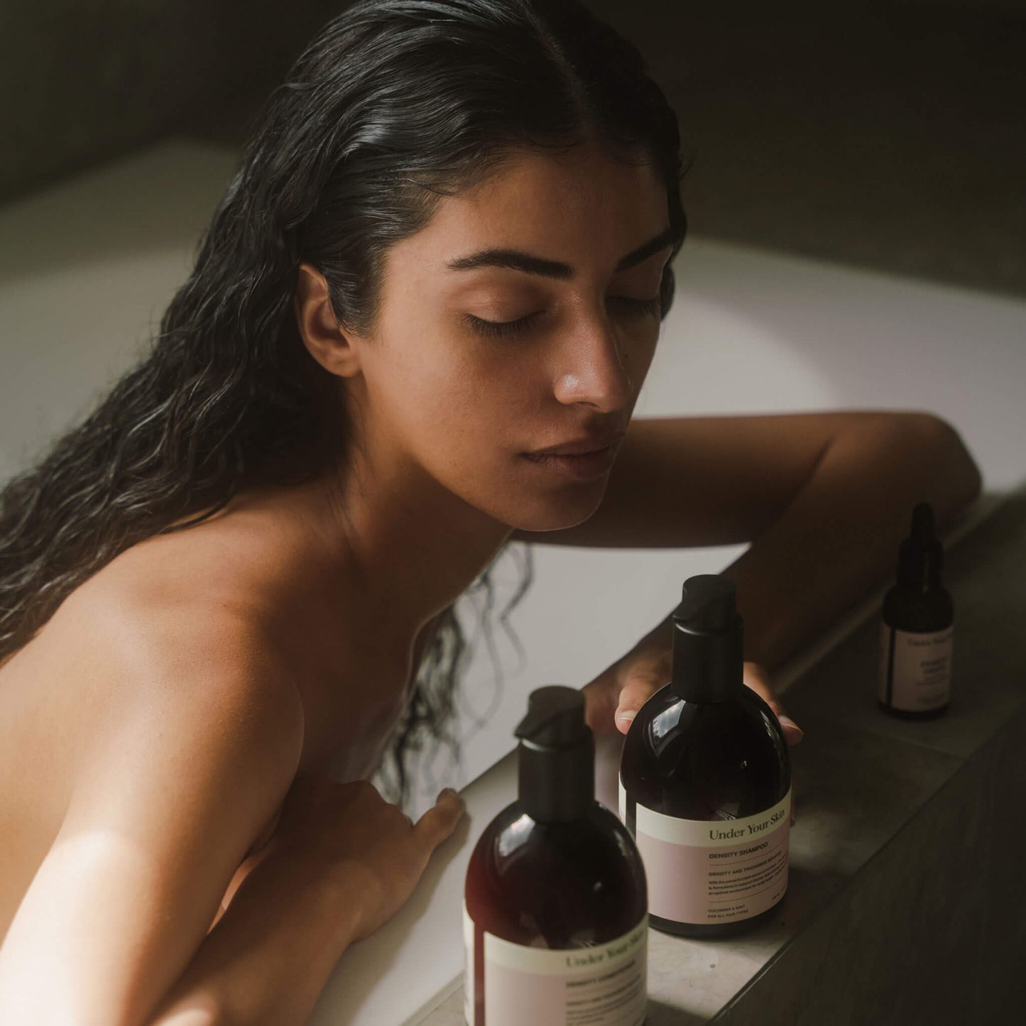 Kvinna med mörkt, långt hår i badkar i badrumsmiljö, lutar händerna på badkarskanten och tittar ner mot Hair Growth Kit-produkterna på kanten.