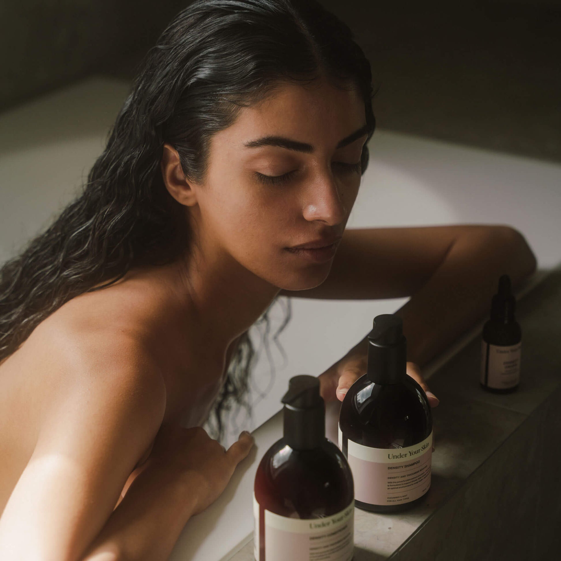 Kvinna med mörkt, långt hår i badkar i badrumsmiljö, lutar händerna på badkarskanten och tittar ner mot Hair Growth Kit-produkterna på kanten.