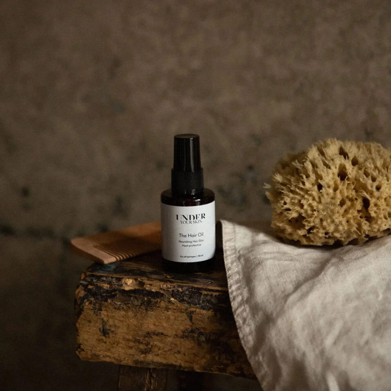 Hårolja på en gammal träbänk tillsammans med en kam och handduk, med mörk gråbeige väggfärg i bakgrunden i badrumsmiljö.