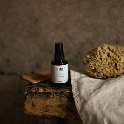 Hårolja på en gammal träbänk tillsammans med en kam och handduk, med mörk gråbeige väggfärg i bakgrunden i badrumsmiljö.