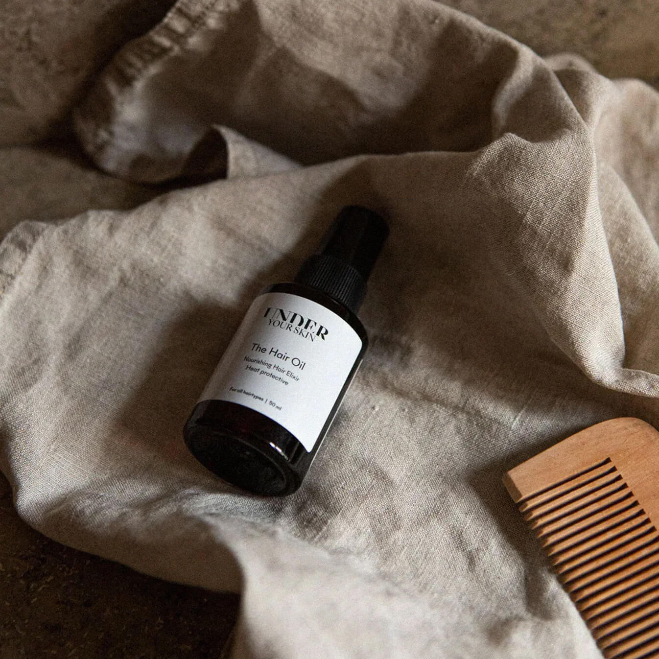 Hårolja liggande på en linnehandduk tillsammans med en träkam bredvid, i badrumsmiljö.