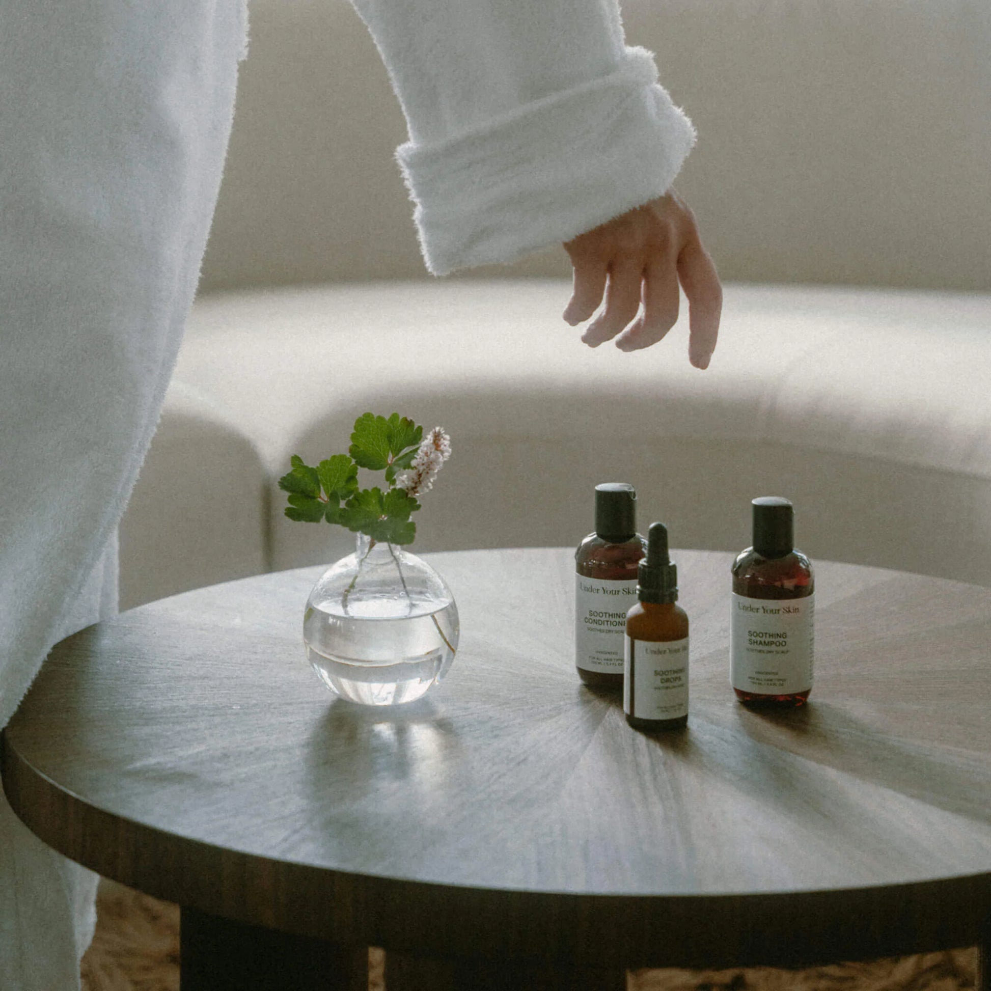 Soothing Kit i reseformat på ett runt träbord, med en kvinna i morgonrock som böjer sig fram för att ta produkten.