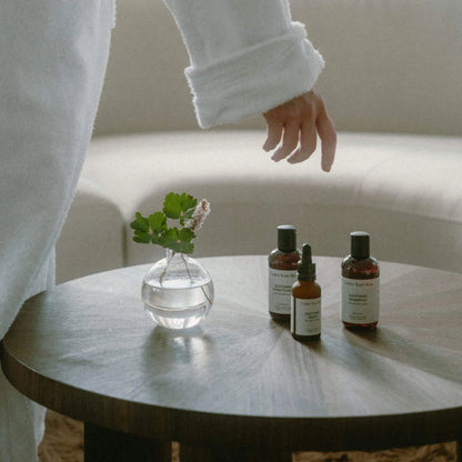 Soothing Kit i reseformat på ett runt träbord, med en kvinna i morgonrock som böjer sig fram för att ta produkten.