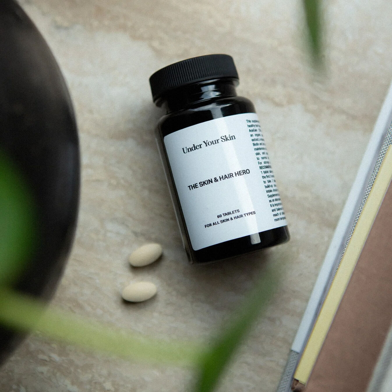 Skin and Hero kosttillskott liggandes på ett bord med några utspridda tabletter och en suddig växt i förgrunden på sidan.