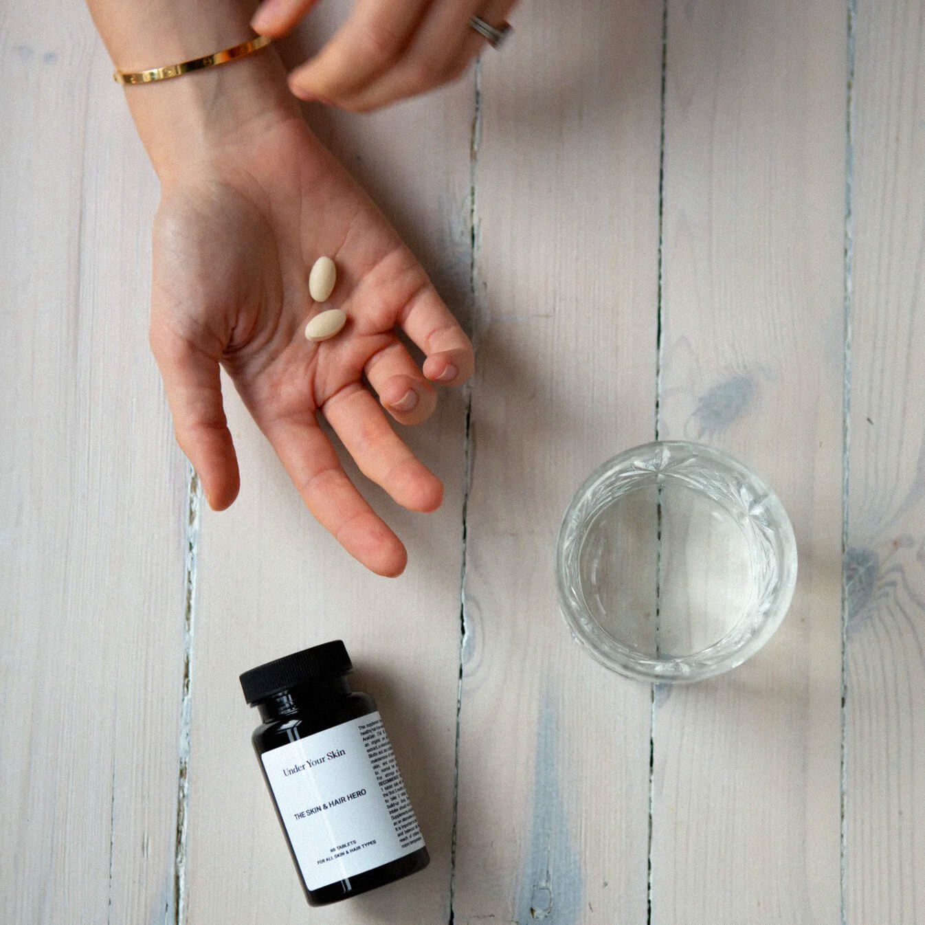 Hand på ett bord tillsammans med två tabletter, ett glas vatten och produkten liggandes på bordet.