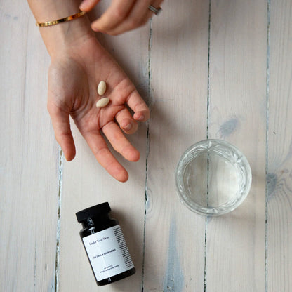 Hand på ett bord tillsammans med två tabletter, ett glas vatten och produkten liggandes på bordet.