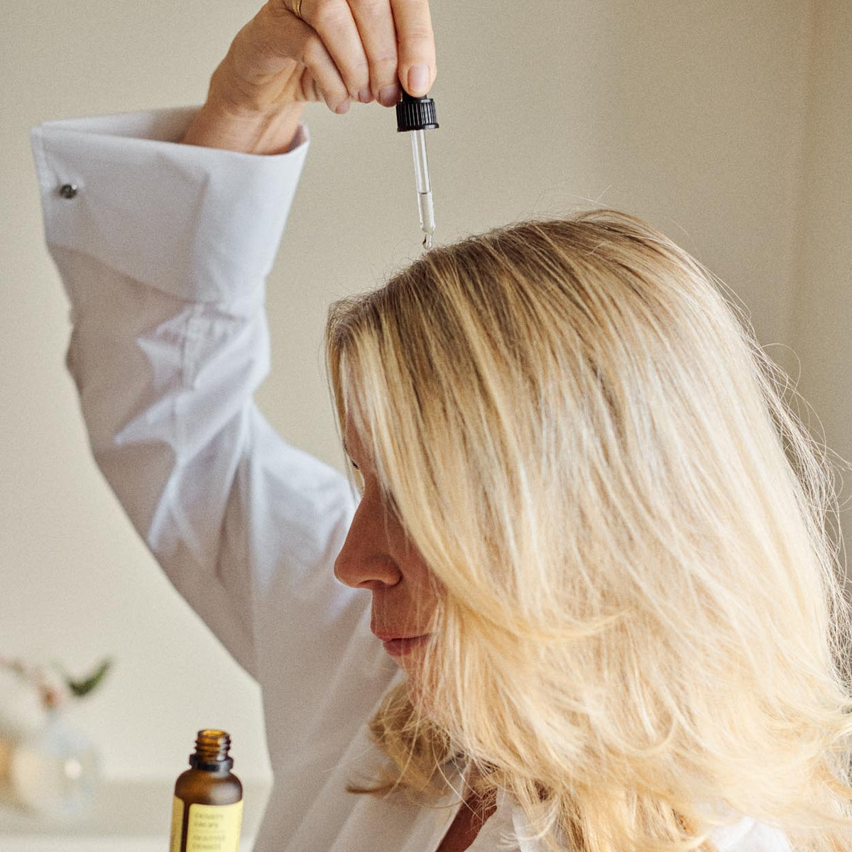 A woman with blonde hair wearing a white shirt holds a small amber dropper bottle in one hand and positions a glass dropper above her hair, suggesting a hair treatment or oil application.