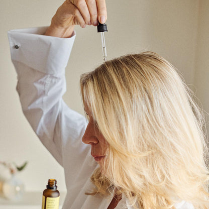 A woman with blonde hair wearing a white shirt holds a small amber dropper bottle in one hand and positions a glass dropper above her hair, suggesting a hair treatment or oil application.