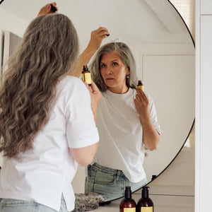 A woman with long gray hair stands in front of a large round mirror, applying product from a small bottle to her hair. Her reflection shows her looking into the mirror, wearing a white t-shirt and light blue jeans.
